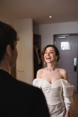 Güzel, genç bir gelin, zarif takılar ve beyaz elbiseler içinde düğün töreninden sonra modern otel odasının kapısında durup gülerken ve damada bakarken, mutlu bir çift. 