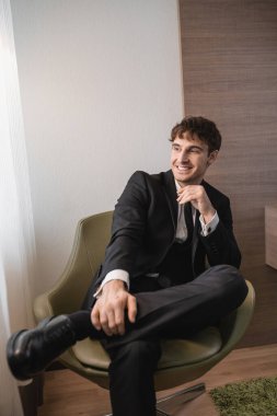 Siyah takım elbiseli, kravatlı ve klasik ayakkabılı mutlu bir adam rahat koltukta oturuyor ve düğün günü gözlerini kaçırıyor, modern otel odasında dinleniyor, yakışıklı bir damat. 