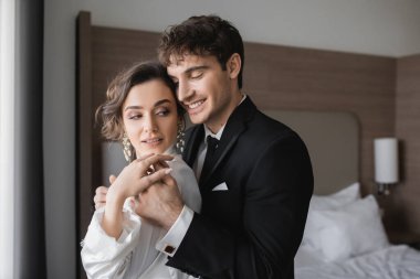 happy groom in classic formal wear touching hand of elegant young bride in jewelry and white dress while standing together in modern hotel room during their honeymoon after wedding  clipart