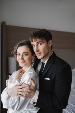 Klasik resmi olarak damat, balayından sonra modern otel odasında birlikte dururken mücevher ve beyaz elbiseli mutlu gelini kucaklıyor. 