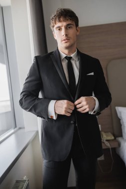 handsome man in classy formal wear with black tie and white shirt buttoning blazer and standing in modern hotel room near window, groom on wedding day, special occasion   clipart