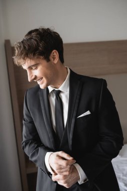 Resmi kıyafetli, şık siyah kravatlı, beyaz gömlekli modern otel odasında gülümseyen, düğün gününde damat olan pozitif bir damat.  