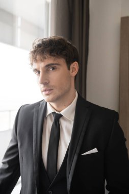 Modern otel odasında dikilen siyah kravatlı ve beyaz gömlekli şık resmi bir damat portresi. Ve pencerenin yanındaki kameraya bakıyor. Düğün gününde damat, özel bir durum.  