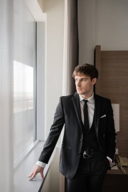 Siyah kravatlı, beyaz gömlekli, şık giyinmiş yakışıklı bir adam. El ele tutuşmuş ve pencerenin yanındaki modern otel odasının pencere eşiğine yaslanmış. Düğün gününde damat. 