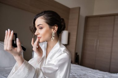 Beyaz ipek bornozlu çekici bir kadın düğün için hazırlanırken dudaklarına dokunuyor, cep aynası tutuyor ve düğün günü otel odasında oturuyor, özel bir gün, genç bir gelin.