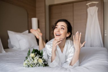 happy woman with brunette hair lying white silk robe and showing engagement ring on finger next to bridal bouquet on bed in hotel room with wedding dress on blurred background, young bride clipart