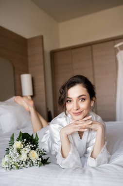 Kumral, beyaz ipek bornozlu, elleri sımsıkı bağlı çekici bir kadın. Düğün günü otel odasındaki buketin yanında, özel bir gün, genç bir gelin.