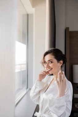 Beyaz ipek bornozlu, heyecanlı genç gelin parmağında nişan yüzüğü düğün günü özel bir gün, dudaklarına yakın bir el için otel takımının içinde pencerenin yanında gülümsediğini gösteriyor. 