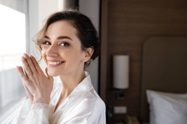 Beyaz ipek bornozla parmağında nişan yüzüğüyle modern otel süitinde kameraya bakan güzel bir genç kadının portresi. Düğün gününde gelin.