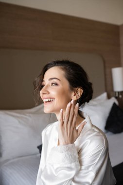 excited young woman with brunette hair in white silk robe showing engagement ring on finger and smiling in hotel room on wedding day, special occasion, young bride clipart