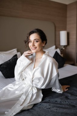 Kahverengi saçlı, gelin makyajlı, çenesine dokunan, rahat bir yatakta beyaz ipek bornozuyla oturan ve düğün günü otel odasındaki kameraya bakan mutlu gelin. 