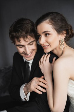 portrait of bride with brunette hair, elegant jewelry and white dress hugging shoulder of happy groom in classic formal wear with tie in modern hotel suite after wedding ceremony  clipart