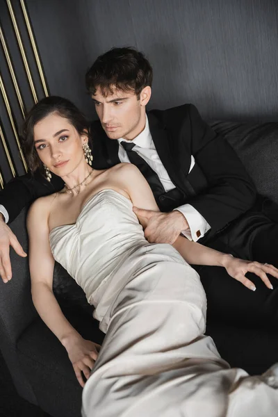 年轻迷人的新娘 戴着华丽的耳环 戴着珍珠 身穿白色婚纱 身穿黑色西服 打着领带 坐在酒店房间里深灰色的沙发上 — 图库照片
