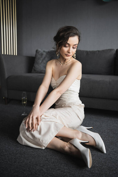 элегантная невеста с брюнетками, сидящая в белом свадебном платье, высокие каблуки и роскошные украшения рядом с бокалом шампанского на полу рядом с диваном в номере отеля 