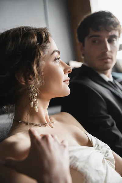 stock image elegant bride in white wedding dress and luxurious jewelry looking away near handsome groom in suit looking at her on blurred background of hotel room 