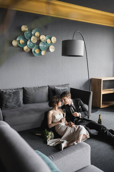 радостная невеста в белом свадебном платье и жених в черном костюме держа бокалы шампанского во время празднования их брака возле свадебного букета после свадьбы в гостиничном номере, счастливые молодожены