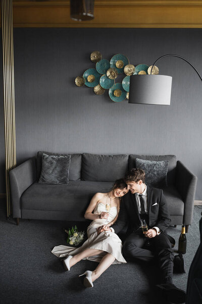 cheerful bride in wedding dress leaning on shoulder of groom in black suit and holding glasses of champagne near bridal bouquet, couch and floor lamp in hotel room 
