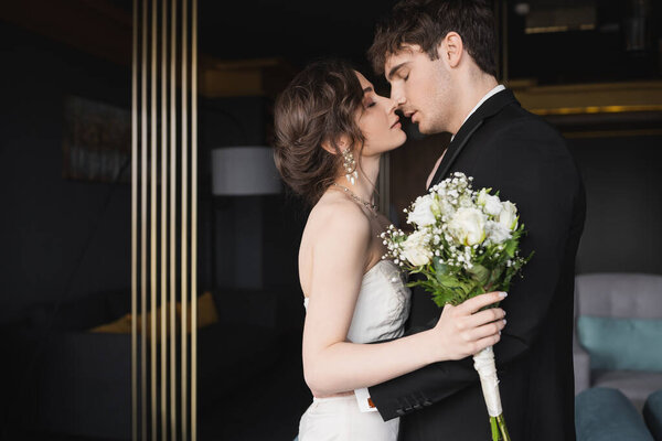side view of good looking groom in black formal wear kissing brunette bride in elegant wedding dress holding bridal bouquet with flowers while standing in hotel lobby 