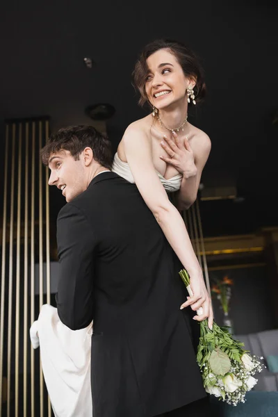 ホテルのロビーに立っている間に花のブライダル花束を保持白いウェディングドレスの化粧で陽気な花嫁を持ち上げる黒の正式な摩耗で良い探して新郎 — ストック写真