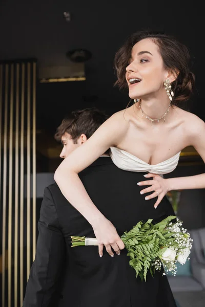 young groom in black formal wear lifting cheerful bride in white wedding dress with opened mouth holding bridal bouquet of flowers while standing in hotel lobby