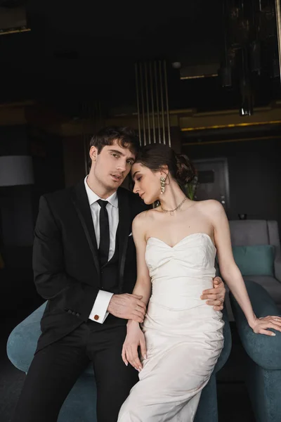 穿着黑色西服的英俊新郎在现代酒店房间里 穿着白色婚纱 头戴珠宝 靠着蓝色沙发 拥抱着迷人的新娘的腰 看着相机 — 图库照片