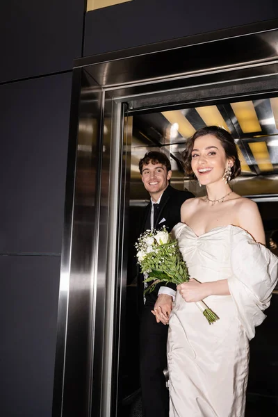 ホテルのエレベーターの外を歩くスーツに花と陽気な新郎の手でブライダル花束を保持白いドレスのかなりの花嫁 幸せな新婚旅行 — ストック写真