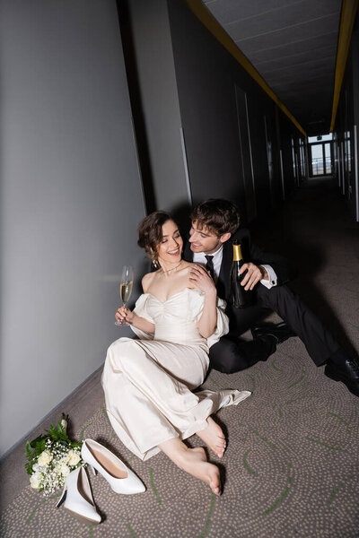 happy groom in black suit holding bottle and sitting near gorgeous bride with glass of champagne next to bridal bouquet and high heels on floor in corridor of modern hotel, newlyweds on honeymoon 