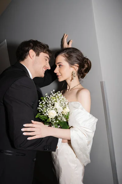 黒いスーツを着た幸せな新郎が壁に寄りかかって花嫁を見る白いウェディングドレスを保持ブライダルブーケを保持しながら一緒に現代ホテルの廊下に立って — ストック写真