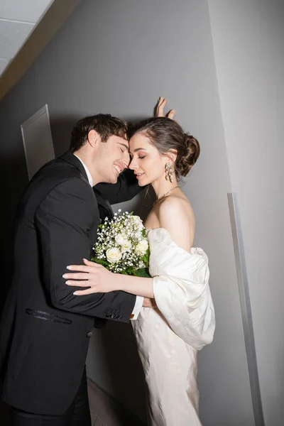 身穿黑色西服的新郎 身穿白色婚纱 头戴婚纱 靠着墙 站在现代宾馆的走廊里 与新婚夫妇度蜜月 — 图库照片