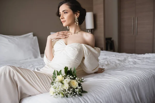 ブライダルブーケの隣のベッドに座って結婚式の日にホテルの部屋でモダンなベッドルームで離れて見る白いドレスと豪華な宝石で魅惑的な花嫁 — ストック写真