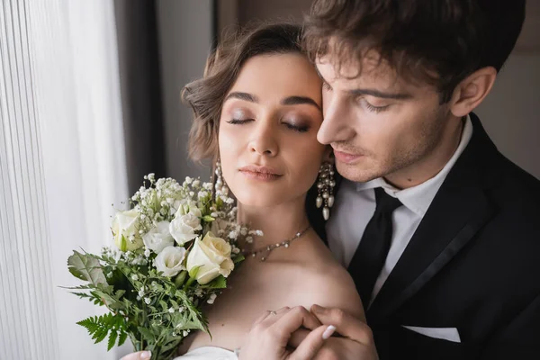 結婚式後の近代的なホテルの部屋で閉じられた目と一緒に立っている間 宝石のかなりの花嫁 古典的な正式な摩耗で新郎と手を保持ブライダル花束と白いドレス — ストック写真