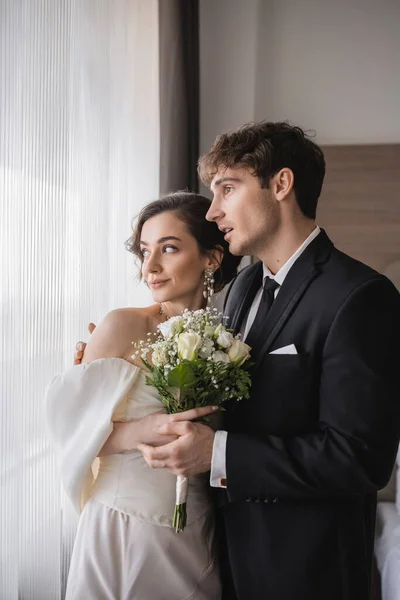 結婚式の後に現代のホテルの部屋でブライダル花束とエレガントな若い花嫁の横に口を開けて立って古典的な正式な摩耗に驚いて新郎 — ストック写真