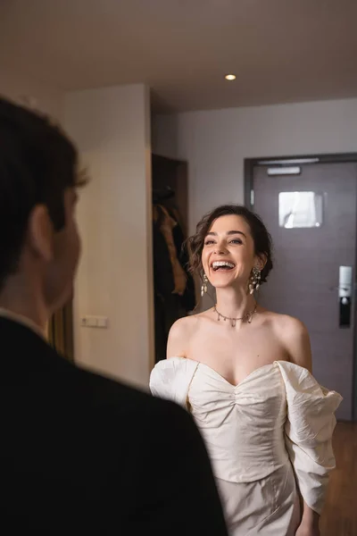 エレガントなジュエリーと白いドレスでかなり若い花嫁は笑っていると結婚式の後 現代のホテルの部屋への入り口のドアの近くに立っている間 ぼやけた新郎を見て 幸せなカップル — ストック写真