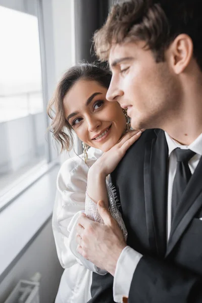 stock image happy bride in jewelry and white silk robe leaning on shoulder of blurred groom in classic black suit while standing together in modern hotel room during honeymoon, newlyweds 