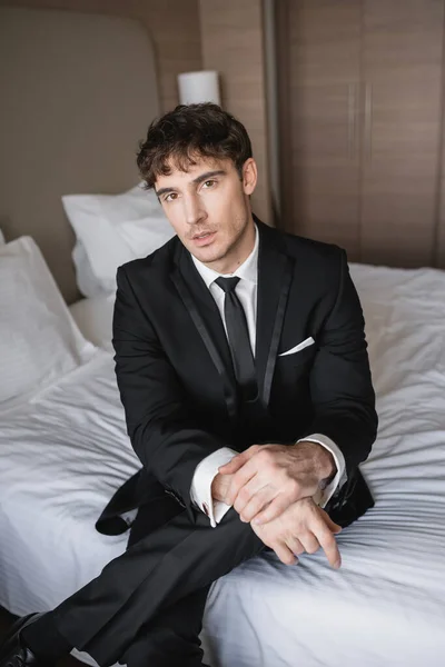 handsome man in classy formal wear with black tie and white shirt sitting on bed in modern hotel room and looking at camera, groom on wedding day, special occasion