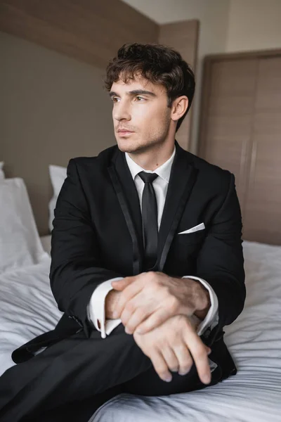 dreamy man in classy formal wear with black tie and white shirt sitting on bed in modern hotel room and looking away, groom on wedding day, special occasion
