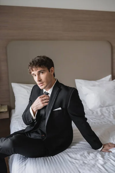 good looking man in classy formal wear with white shirt adjusting black tie while looking at camera and sitting on bed in modern hotel room, groom on wedding day, special occasion