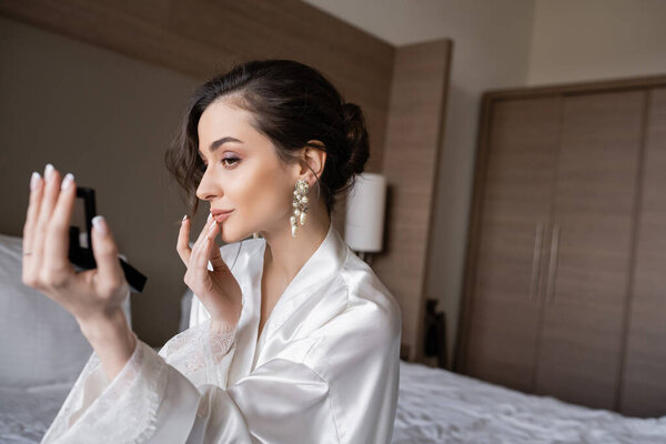 attractive woman in white silk robe preparing for her wedding while touching lips, holding pocket mirror and sitting on bed in hotel room on wedding day, special occasion, young bride