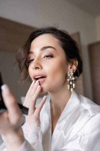 young bride with brunette hair in white silk robe preparing for her wedding while touching lips, holding pocket mirror in hotel room on wedding day, special occasion, 