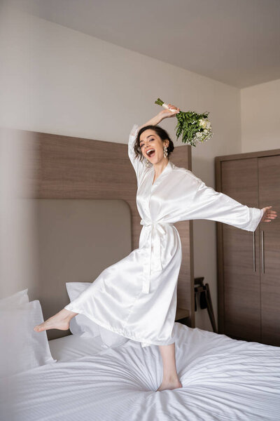 excited young bride with brunette hair in white silk robe and pearl earrings holding bridal bouquet while jumping on bed in hotel room, special occasion, happy woman 