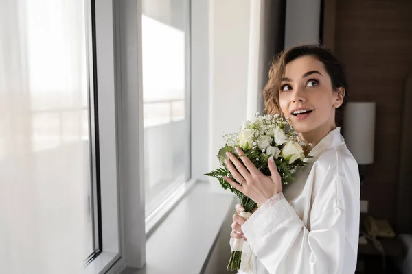 amazed young woman with engagement ring on finger standing in white silk robe and holding bridal bouquet while looking up next to window in hotel suite, special occasion, bride on wedding day