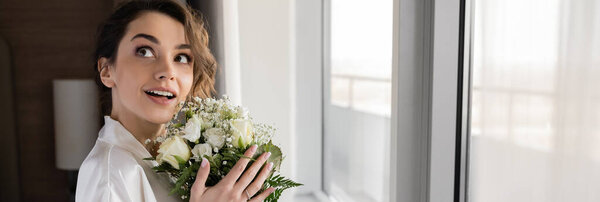 amazed woman with engagement ring on finger standing in white silk robe and holding bridal bouquet while looking up next to window in hotel suite, special occasion, bride on wedding day, banner 