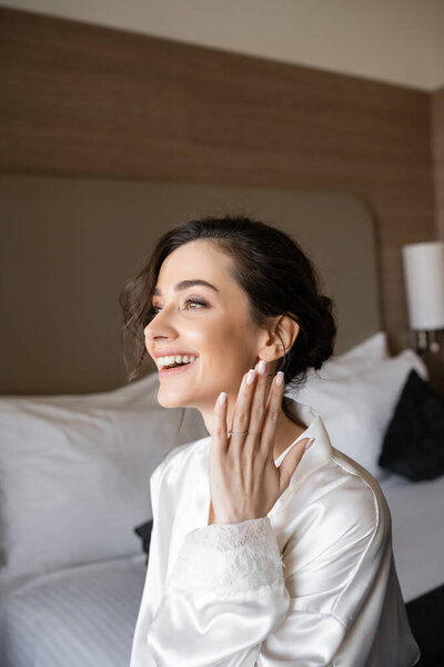 excited young woman with brunette hair in white silk robe showing engagement ring on finger and smiling in hotel room on wedding day, special occasion, young bride