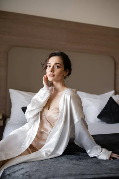 stock image delightful young bride with brunette hair and bridal makeup touching her face and sitting in white silk robe on comfortable bed of hotel suite on wedding day 