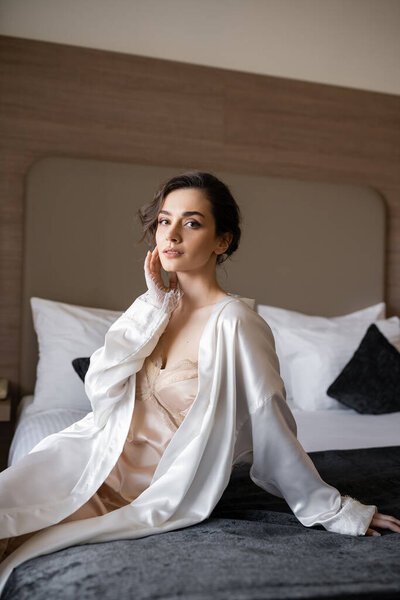 delightful young bride with brunette hair and bridal makeup touching her face and sitting in white silk robe on comfortable bed of hotel suite on wedding day 