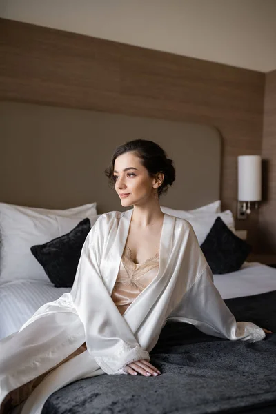 charming bride with brunette hair and bridal makeup sitting in white silk robe on comfortable bed and looking away in hotel suite before her wedding, special occasion