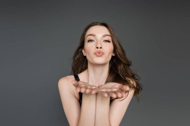 Pretty and young brunette model with hairstyle and natural makeup looking at camera while blowing air kiss while standing and posing isolated on grey,  flirt, chic  clipart