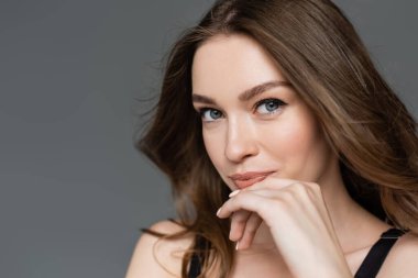 Close up view of confident and pretty young model with everyday makeup and brunette hair looking at camera and holding hand near face isolated on grey, portrait  clipart