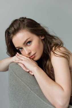 Portrait of pretty and young brunette woman with natural makeup and hairstyle looking at camera while relaxing on grey modern armchair isolated on grey   clipart