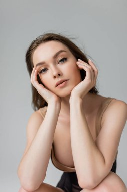 Portrait of young and seductive woman with brunette hair and natural makeup wearing bodysuit while touching face and looking at camera isolated on grey  clipart